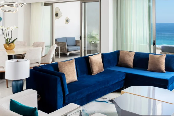 Modern living room with a blue sectional sofa, dining area, and ocean-view balcony. Decor includes a white orchid and abstract wall art.