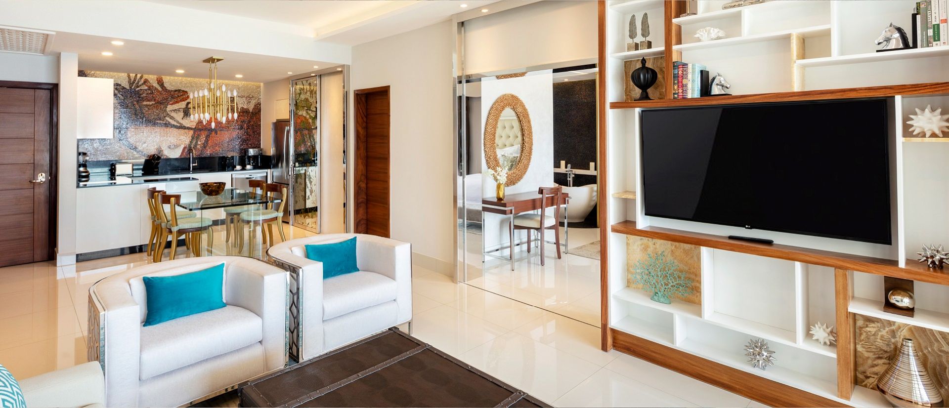 Modern living room with white sofas, turquoise cushions, large TV, and glass dining area. Open kitchen with mosaic wall art in the background.