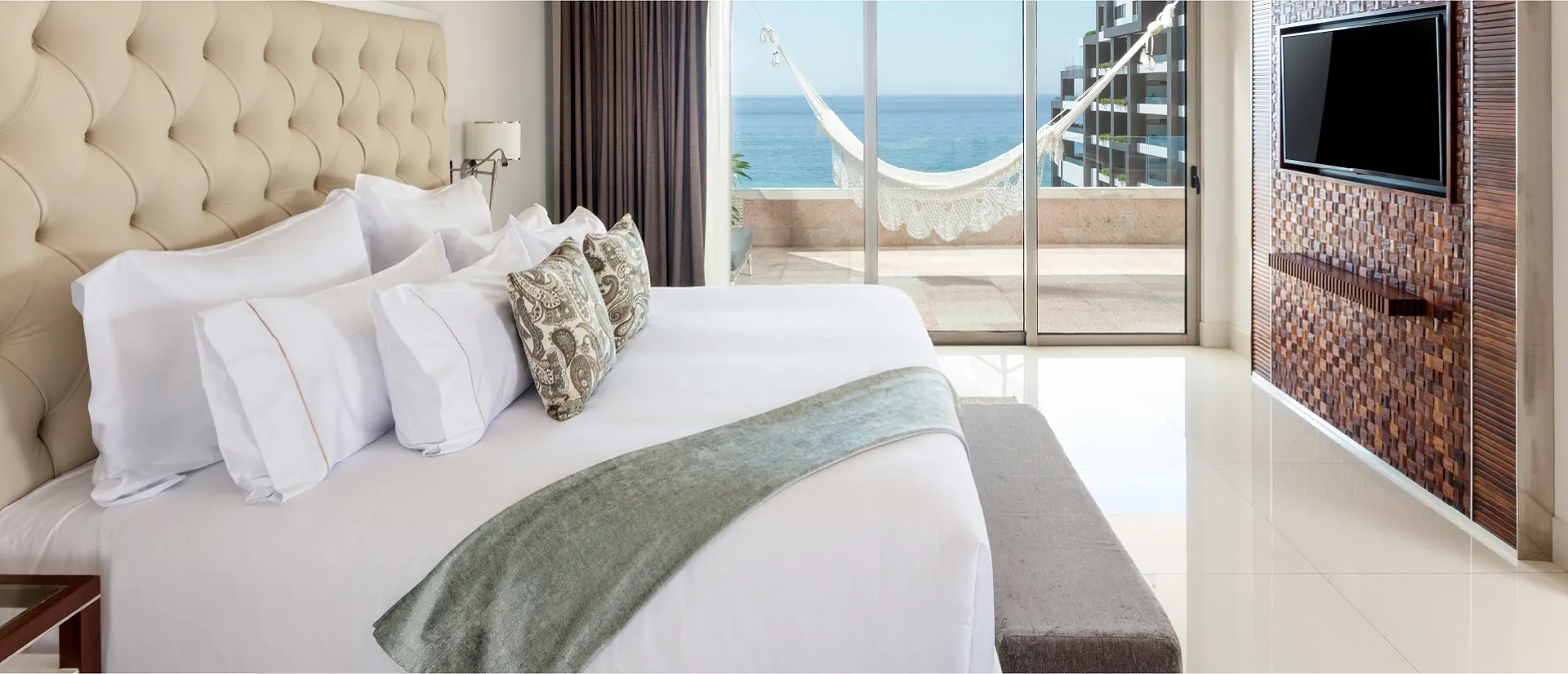 Luxurious bedroom with a large bed, decorative pillows, and a view of the ocean through glass doors leading to a balcony with a hammock.