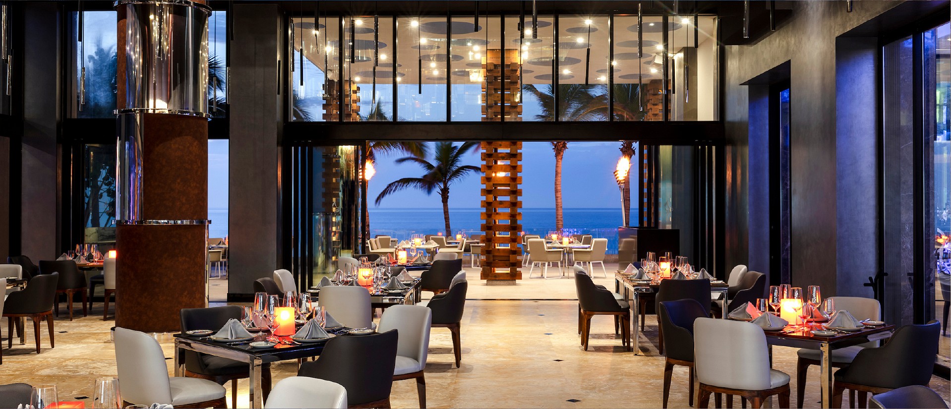 Elegant restaurant with modern decor, set tables, and large windows offering a view of palm trees and the ocean at dusk.