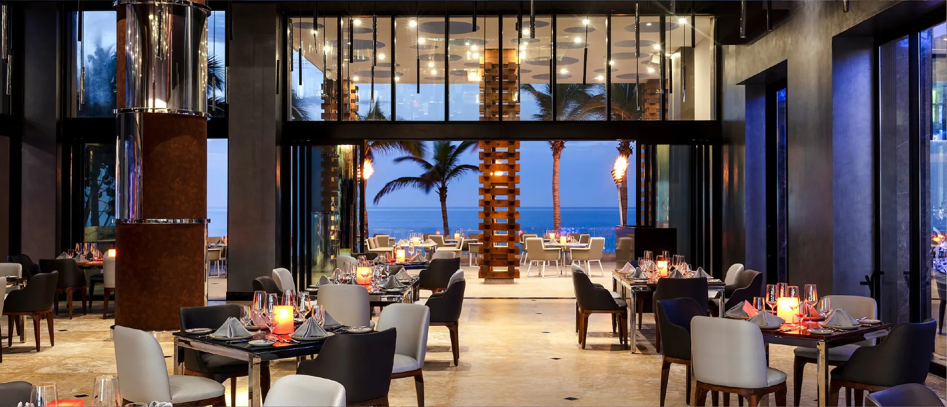 Elegant restaurant with modern decor, set tables, and large windows offering a view of palm trees and the ocean at dusk.