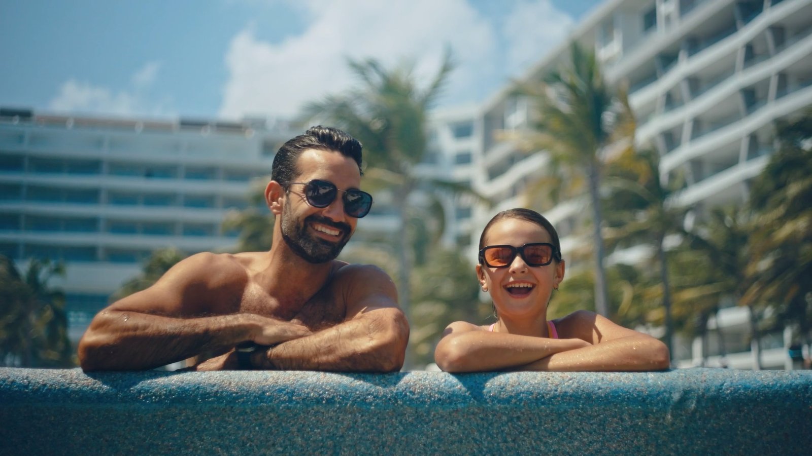 A man and woman in sunglasses smile while leaning on the edge of a pool, with palm trees and a modern building in the background.