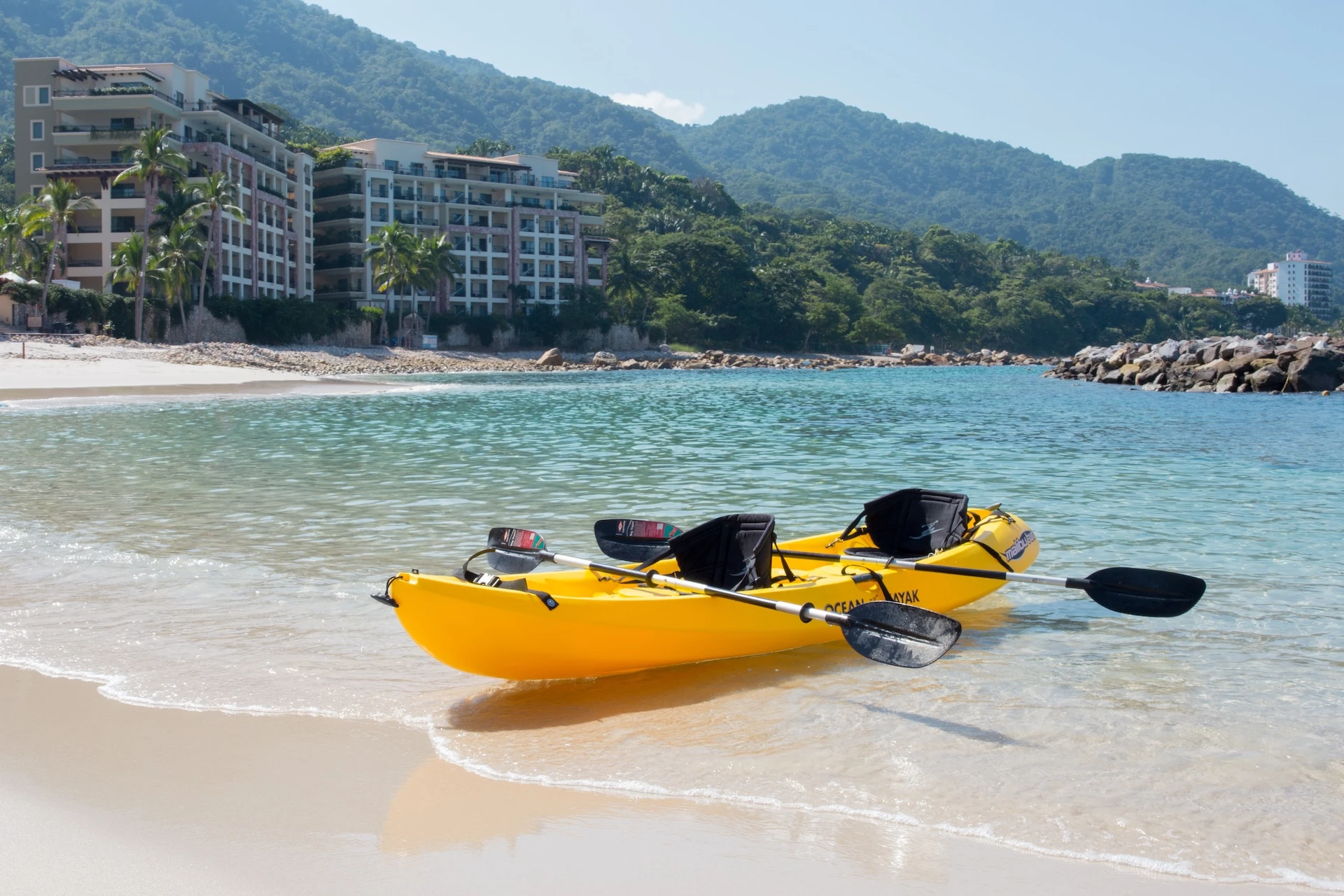 Bright yellow double kayak on Garza Blanca&rsquo;s sandy beach, turquoise water, palm trees, and upscale resort buildings with green hills behind.