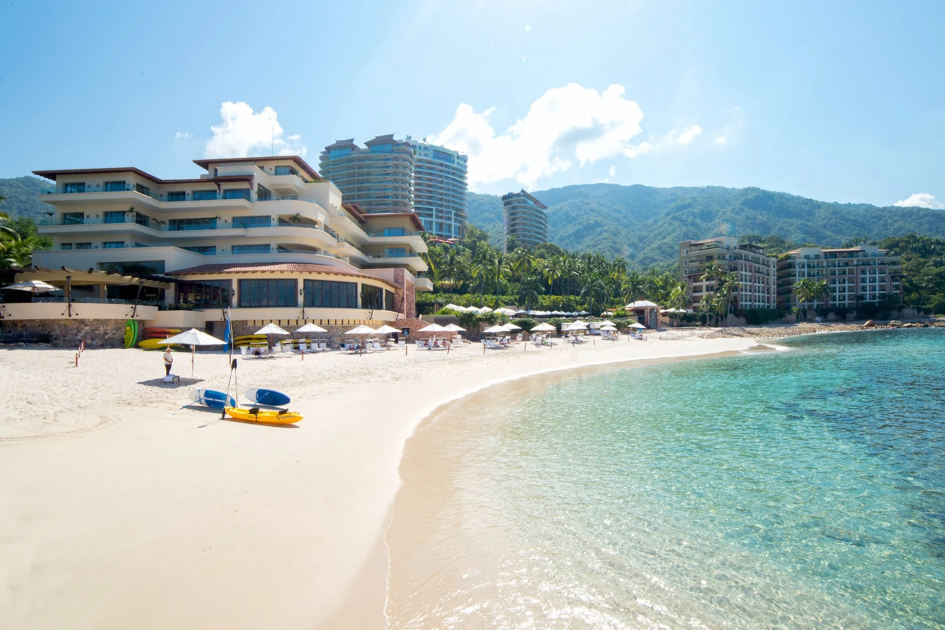 Luxury beachfront resort with white-sand beach, turquoise water, palm trees, kayaks on shore, and modern villa buildings against green mountains.