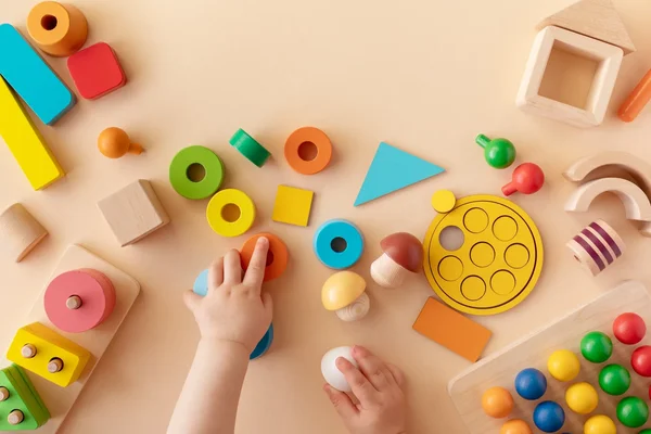 Baby's hands playing with colorful educational wooden blocks and geometric shapes on a light beige surface.