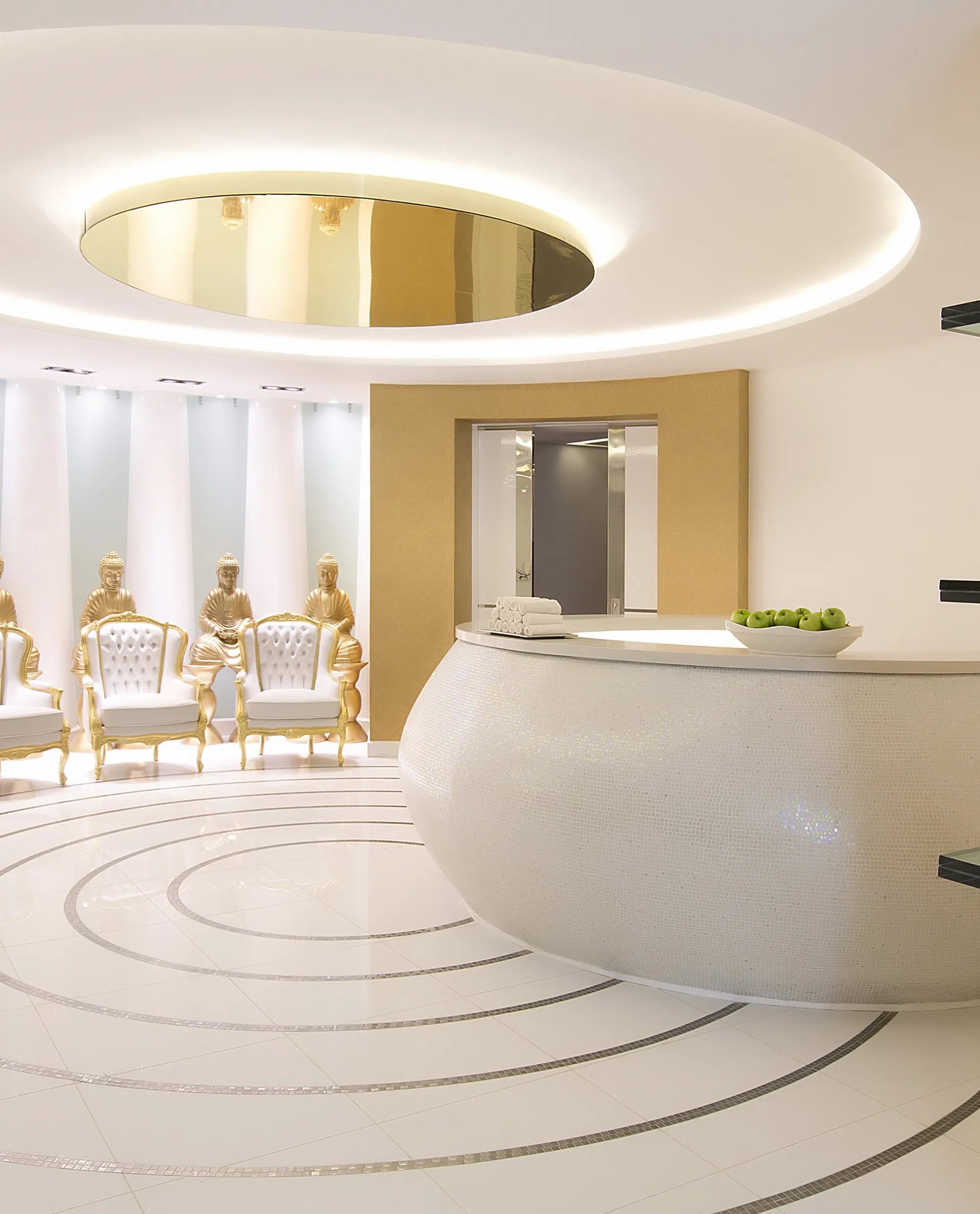 Modern lobby with a circular reception desk, gold accents, decorative vases, and a row of elegant chairs. Ceiling features a large round light.