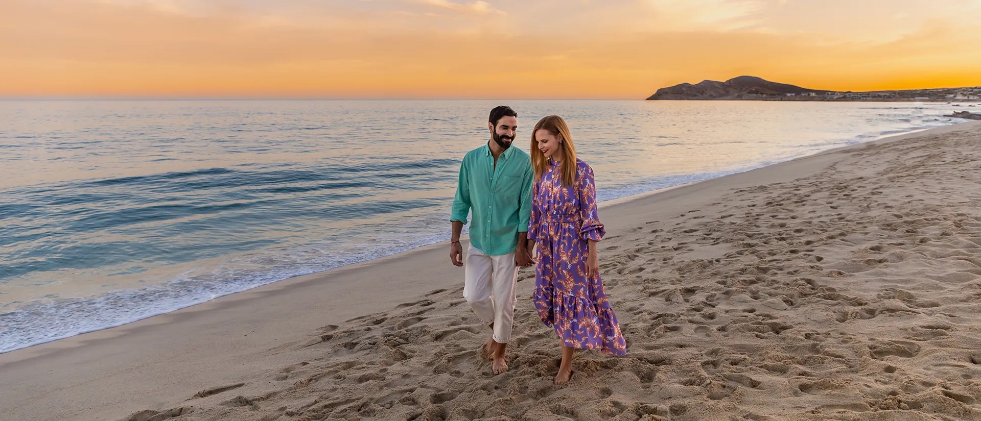 A couple walks barefoot along a sandy beach at sunset, with gentle waves and an orange sky in the background.