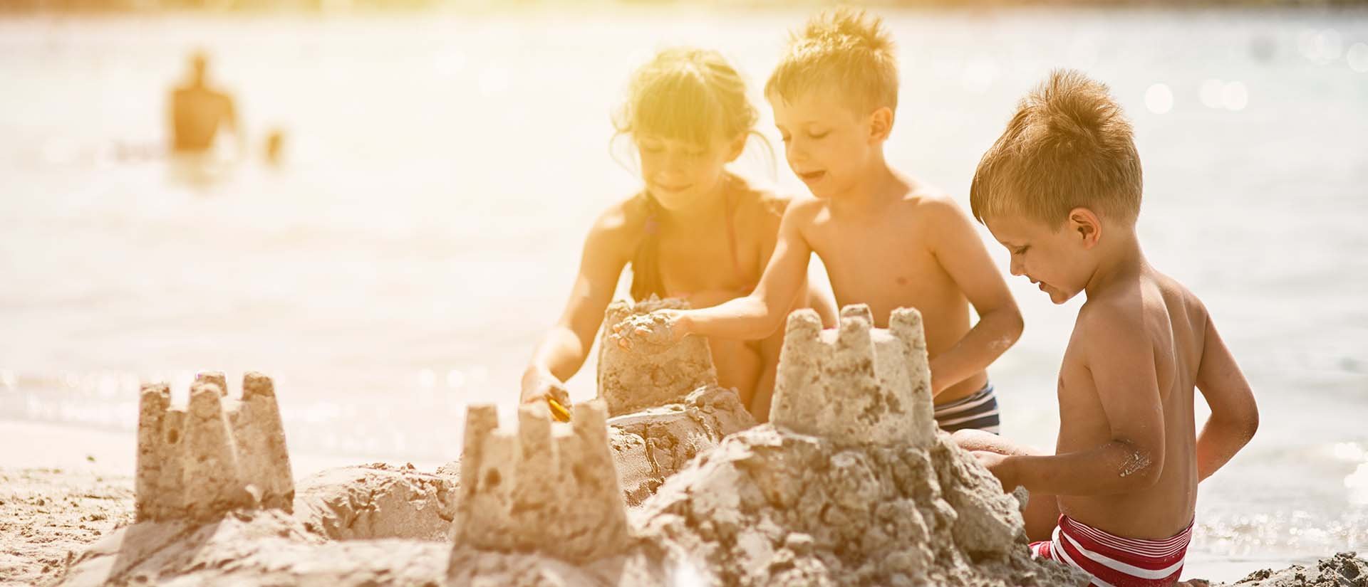 Three children in swimsuits build sandcastles on a sunny beach, with the ocean in the background.