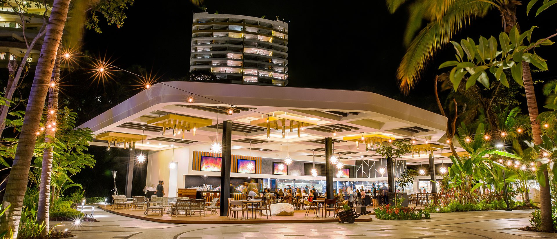 Outdoor restaurant at night with modern design, surrounded by tropical plants and illuminated by string lights, with a tall building in the background.