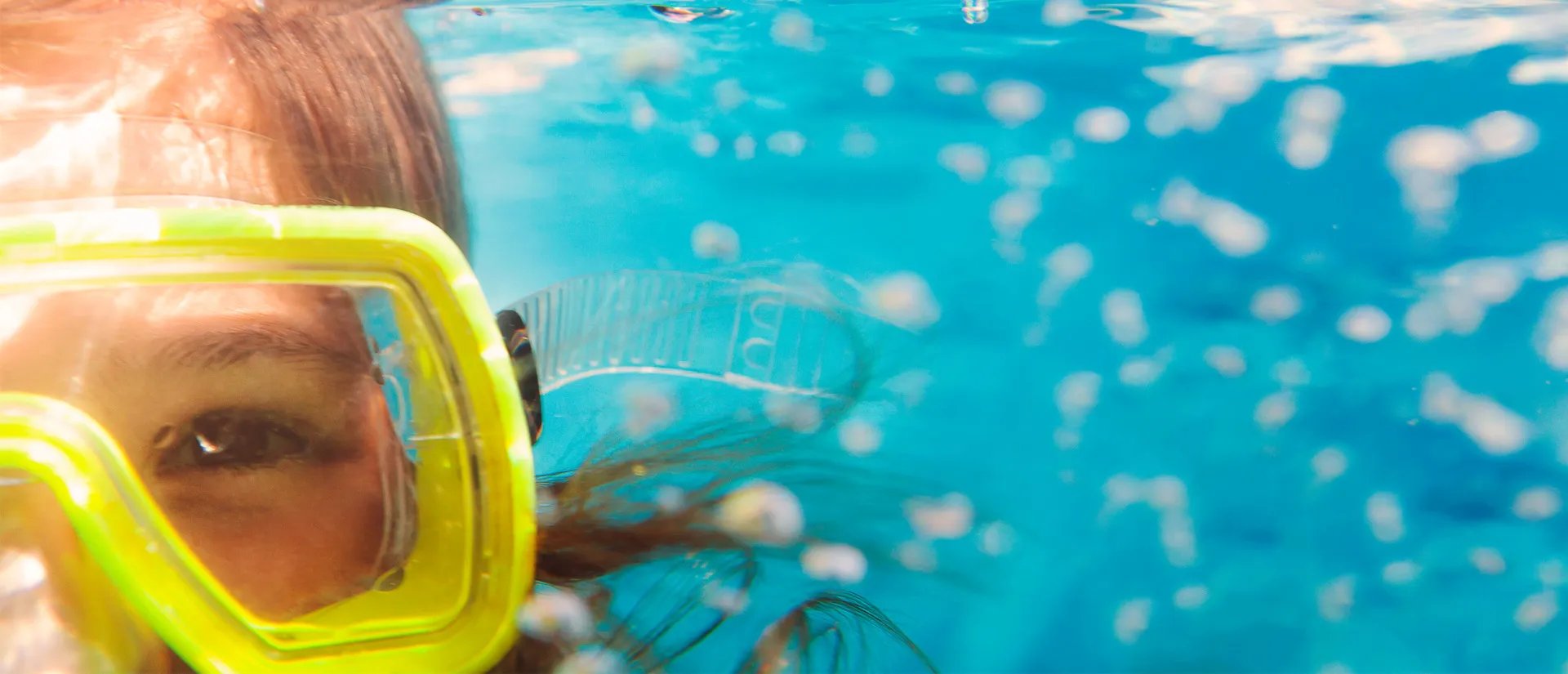 Close-up of a person wearing a yellow snorkel mask, partially submerged in clear blue water with bubbles surrounding them.