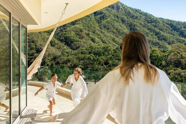 A woman in a robe opens her arms to two children running toward her on a balcony with mountain views and a hammock.