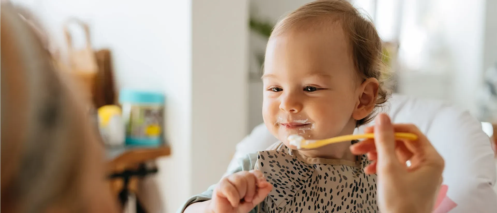 Joyful baby with a patterned bib being spoon-fed yogurt; cream around mouth in a cozy, bright dining setting.