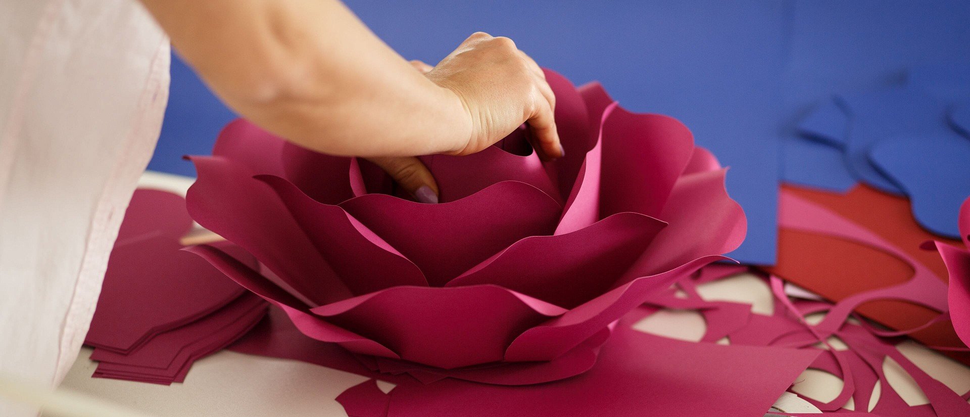 Person assembling a large, pink paper flower with layered petals on a table, surrounded by blue and red paper cutouts.