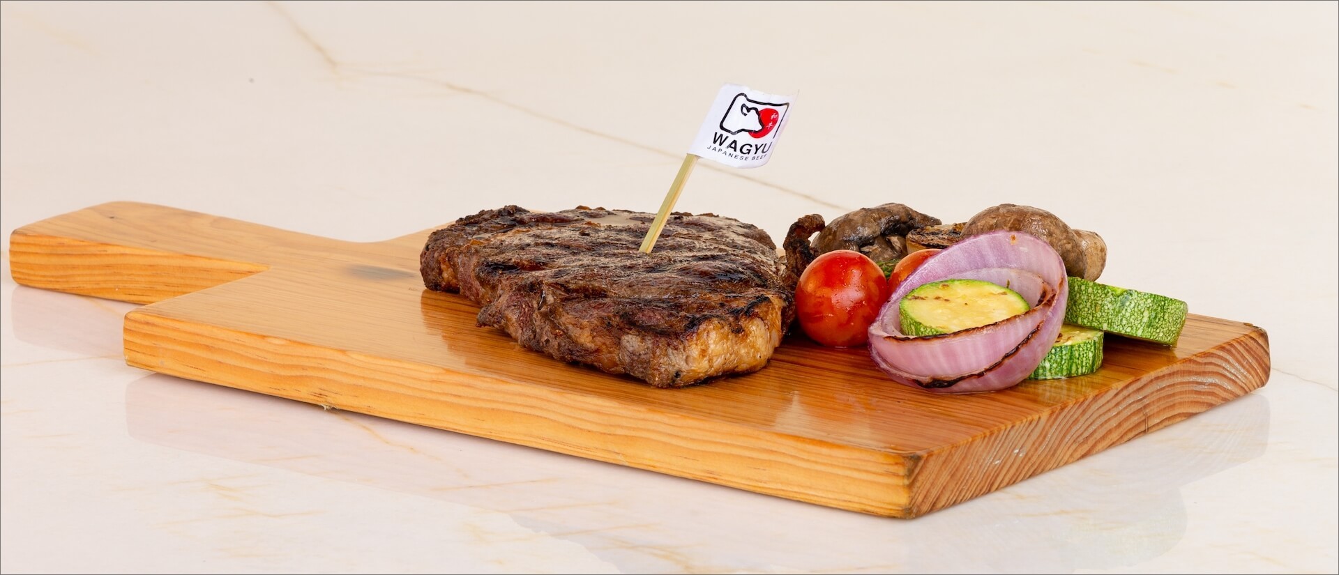 Grilled steak on a wooden board with zucchini, onion, mushroom, and tomato, garnished with a small flag.