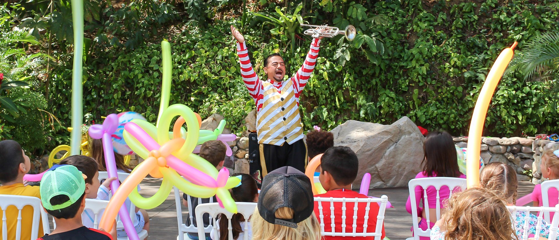 A performer in a colorful outfit raises a trumpet, entertaining a seated audience of children surrounded by colorful balloon animals.