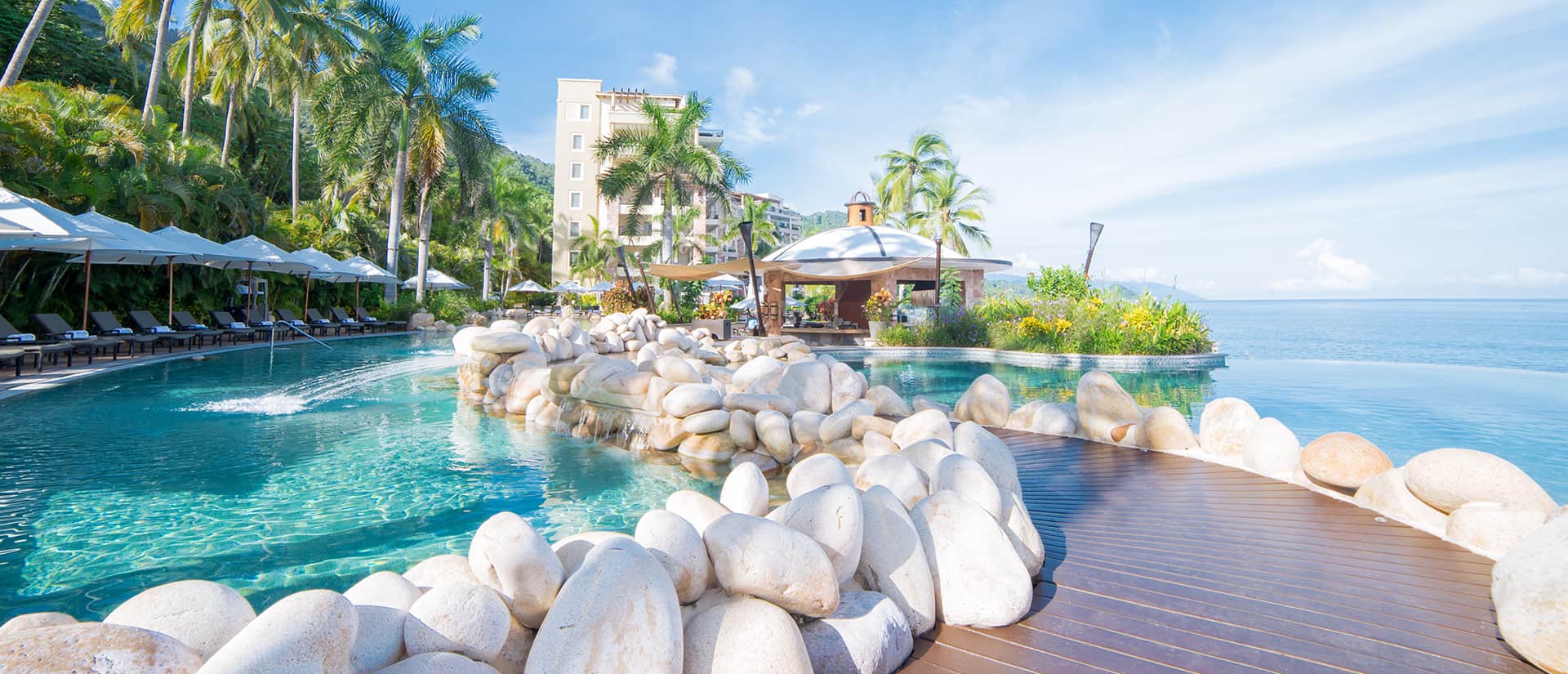 Luxurious resort pool with stone accents, surrounded by palm trees and lounge chairs, overlooking a calm ocean under a clear blue sky.