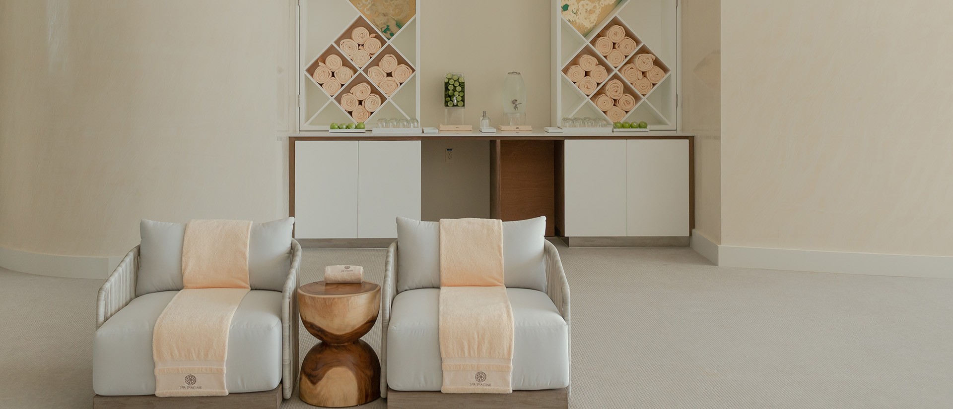 Minimalist spa room with two cushioned lounge chairs, a small wooden table, and a counter with towels and decor on shelves.