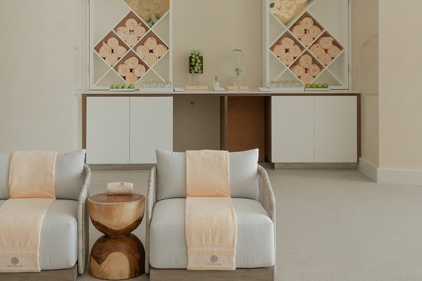Minimalist spa room with two cushioned lounge chairs, a small wooden table, and a counter with towels and decor on shelves.