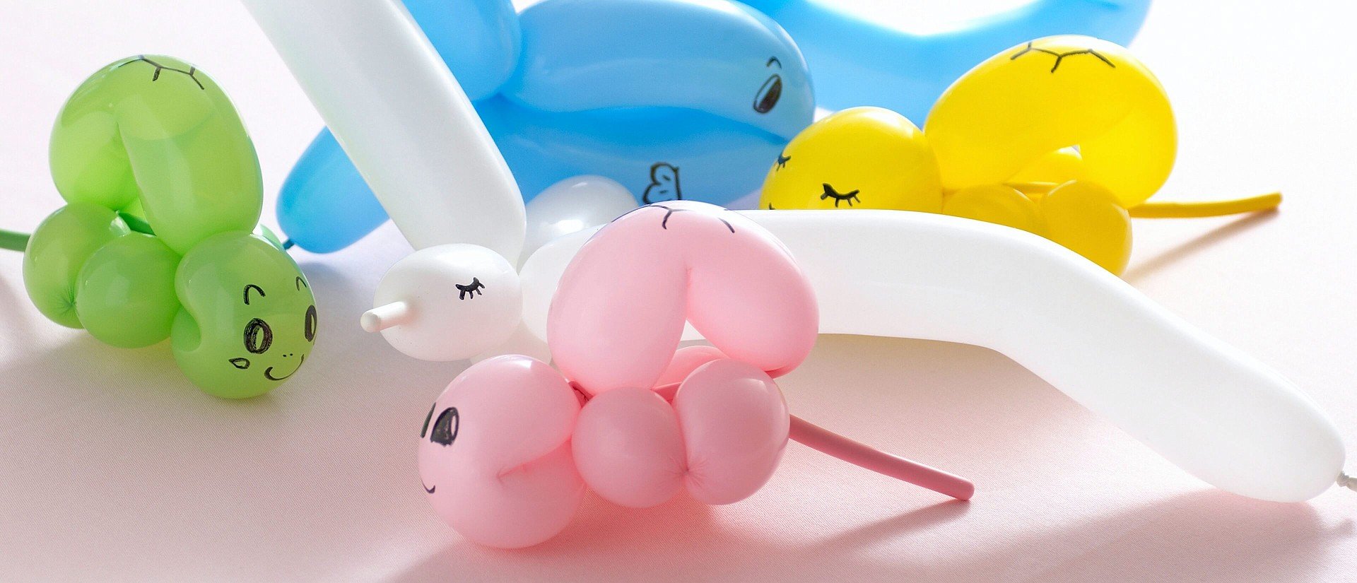 Colorful balloon animals, including a pink unicorn, blue dog, and yellow duck, with drawn faces, on a light pink surface.