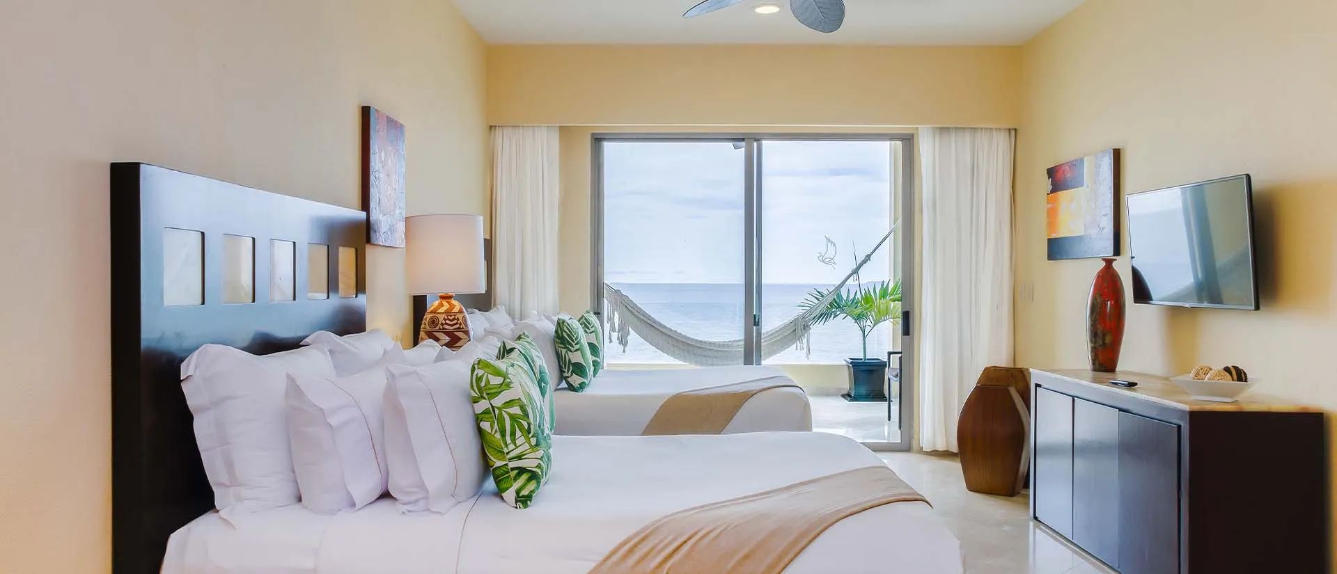 Hotel room with two beds, tropical-themed pillows, a TV, and a balcony with a hammock overlooking the ocean. Bright and inviting decor.