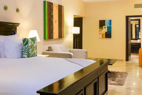 Spacious hotel room with a large bed, tropical-themed pillows, modern artwork, a cozy armchair, and a view into a bathroom.