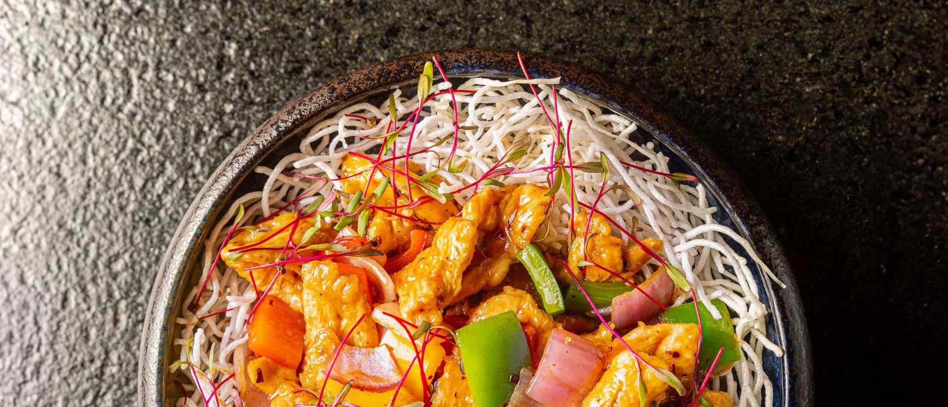 A bowl of stir-fried chicken with colorful bell peppers, onions, and red chili strands on a bed of crispy noodles.