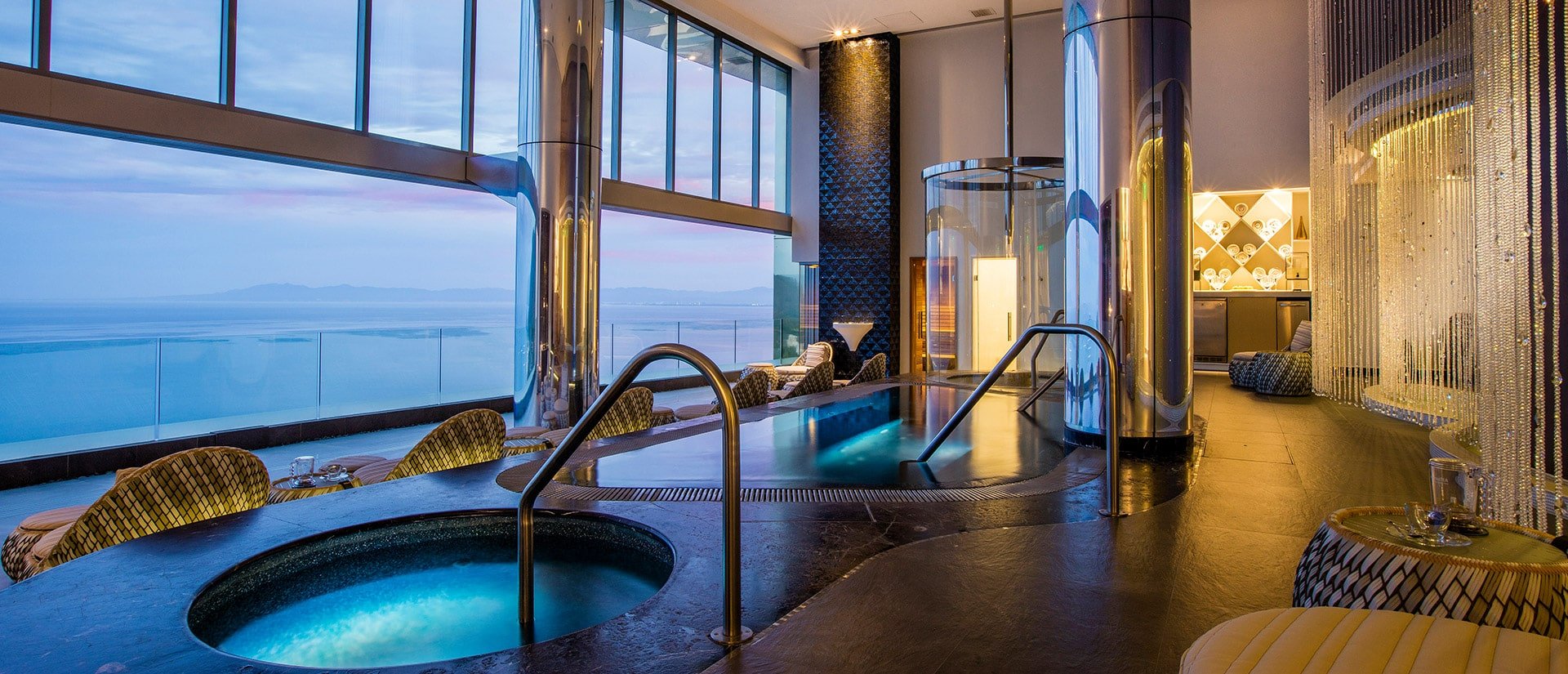 Luxurious spa with indoor hot tubs, modern decor, and large windows offering a panoramic ocean view at sunset.