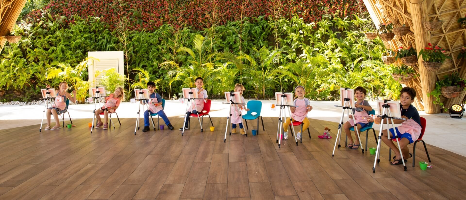 Children painting on canvases in a bright, open space with lush greenery in the background. They are seated in a row, each focused on their artwork.