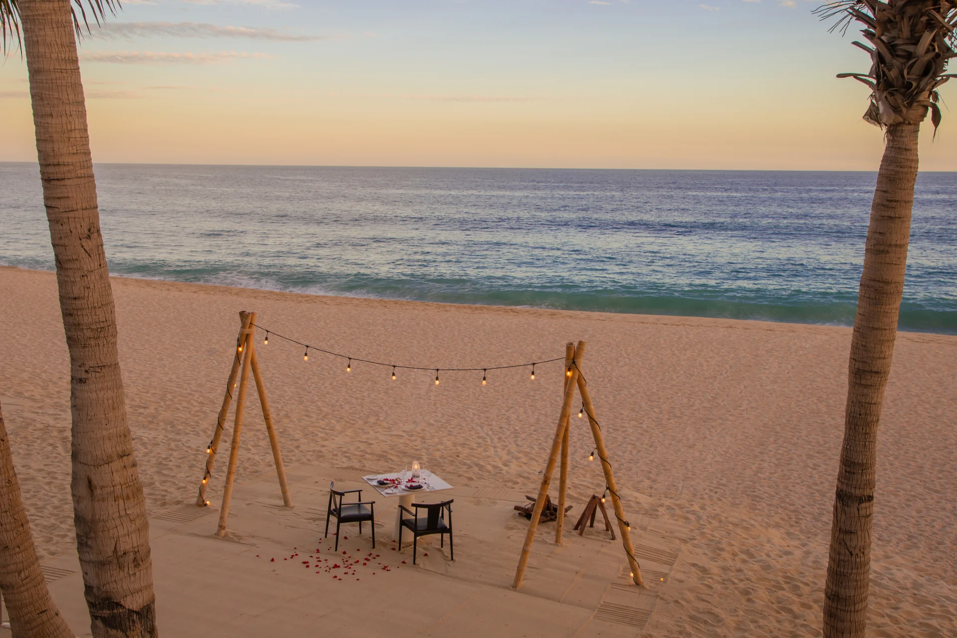 Cena romántica en la playa al atardecer con series de luces, rodeada de palmeras y frente al mar en calma en Garza Blanca Cabo