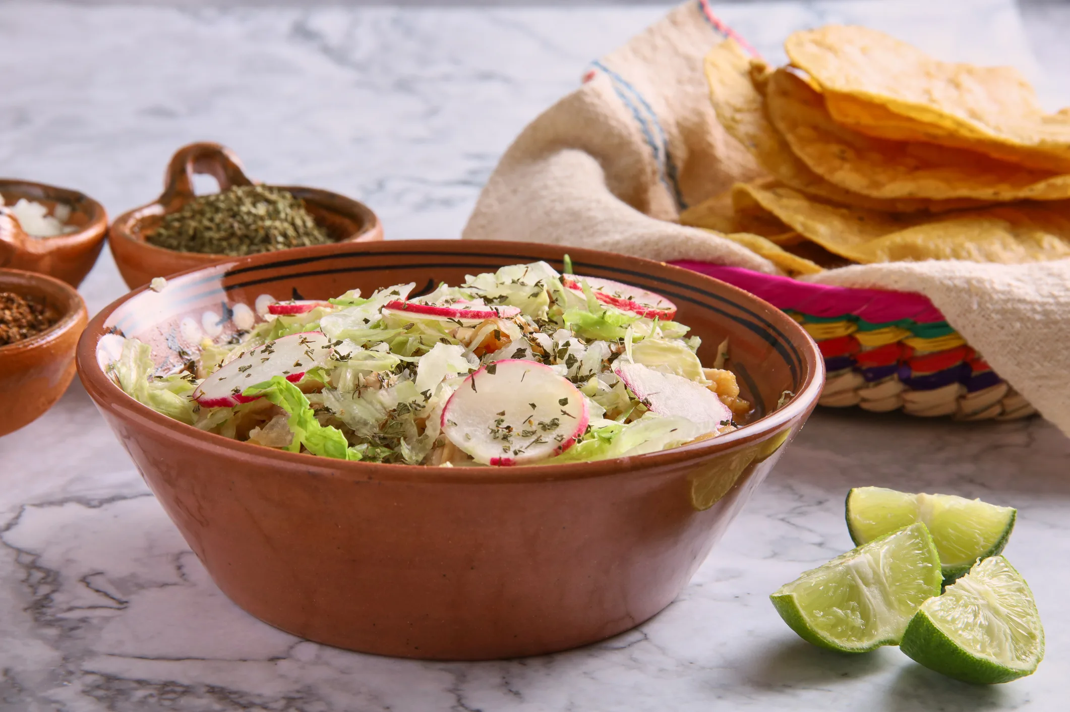 A bowl of salad with radishes and herbs, surrounded by lime wedges, crispy tortillas, and small bowls of spices on a marble surface.