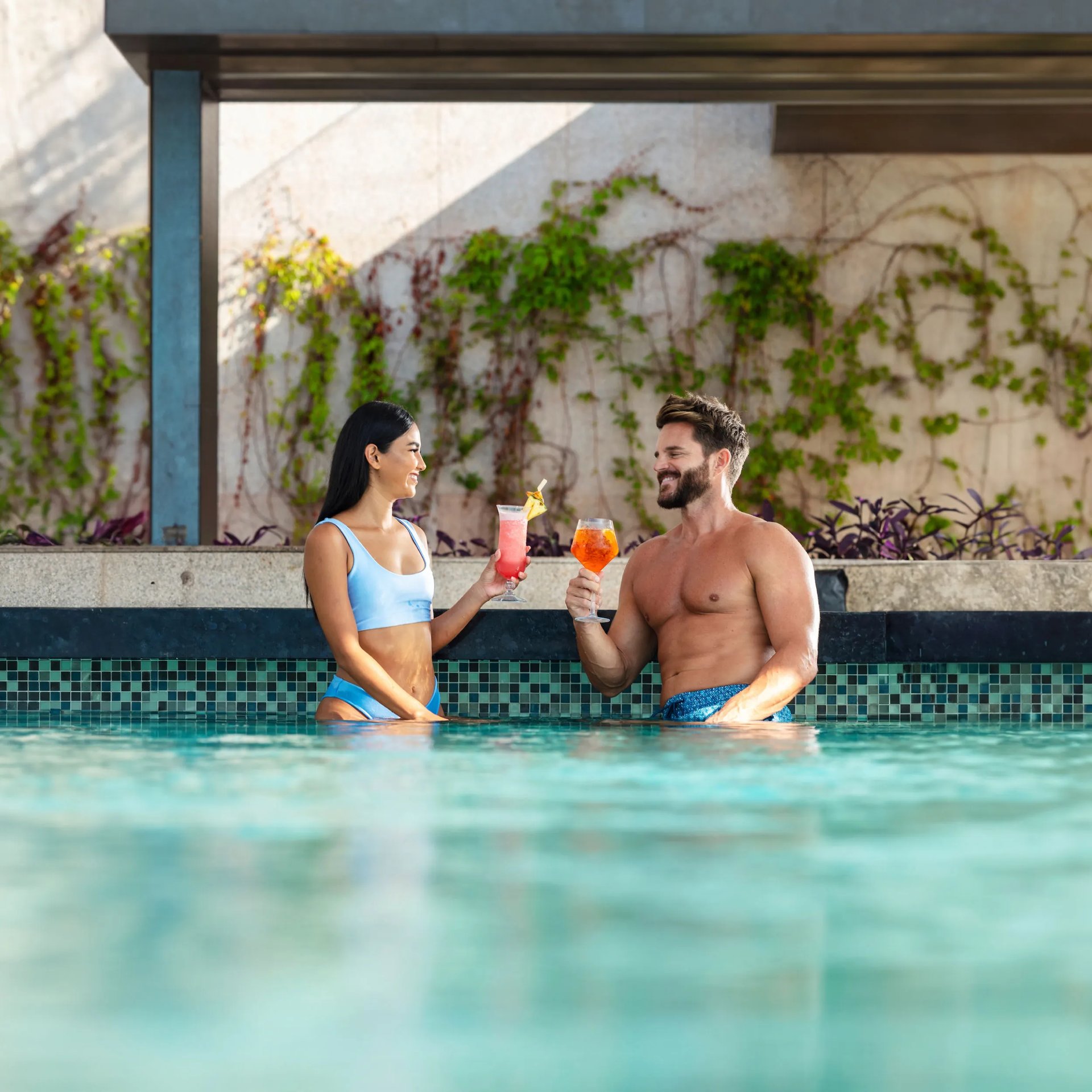 Couple in blue swimsuits enjoy colorful cocktails at a pool edge with lush vines behind Garza Blanca, a luxury all-inclusive resort in Mexico.