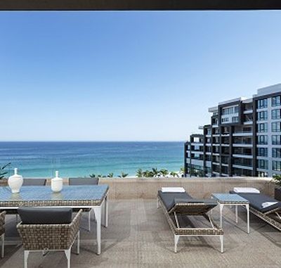 Rooftop terrace with wicker furniture overlooking the ocean, adjacent to modern apartment buildings under a clear blue sky.