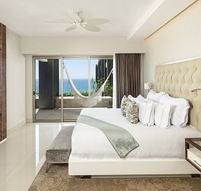 Modern bedroom with a large bed, decorative pillows, and a light throw. Glass doors open to a balcony with a hammock and ocean view.