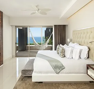 Modern bedroom with a large bed, decorative pillows, and a light throw. Glass doors open to a balcony with a hammock and ocean view.