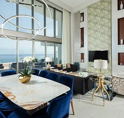 Modern living room with ocean view, featuring a marble dining table, blue chairs, sleek sofas, large windows, and decorative lighting.