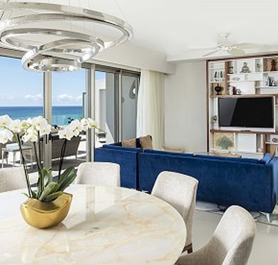 Modern living room with a marble table, gold vase with orchids, blue sofa, wall-mounted TV, and large windows overlooking the ocean.