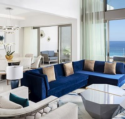 Modern living room with a blue sectional sofa, beige pillows, glass coffee table, and ocean view through large windows. Elegant dining area adjacent.