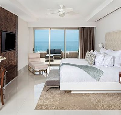 Elegant hotel room with a large bed, TV, and seating area. Glass doors open to a balcony with ocean views. Neutral tones and modern decor.