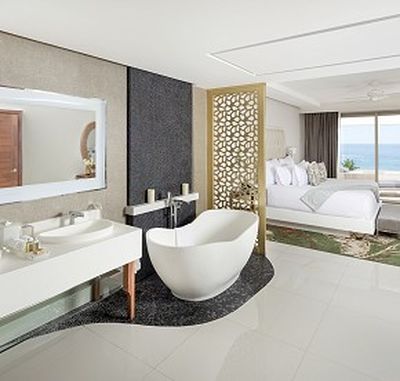 Luxurious hotel suite with a freestanding bathtub, double sink vanity, decorative partition, and a view of the ocean through large windows.