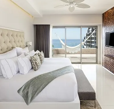 Modern hotel room with a king-size bed, decorative pillows, and a hammock on the balcony overlooking the ocean. Flat-screen TV on the wall.