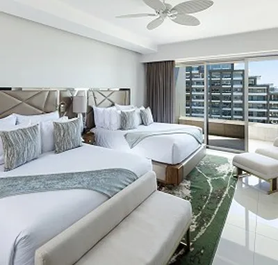 Modern hotel room with two large beds, decorative pillows, a ceiling fan, and a balcony view of city buildings. Bright and elegant decor.
