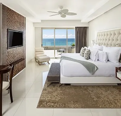 Modern hotel room with ocean view, featuring a large bed, wall-mounted TV, armchair, and sliding glass doors leading to a balcony.