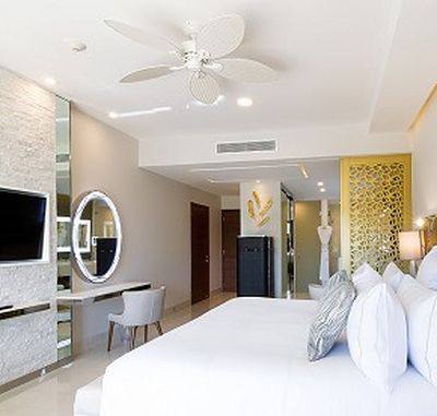 Modern hotel room with a large bed, wall-mounted TV, round mirror, and ceiling fan. Bright decor with neutral tones and decorative screen.