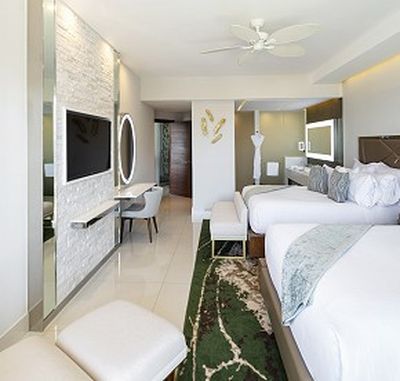 Modern hotel room with two beds, a wall-mounted TV, desk with mirror, and a green patterned rug. Decor includes a ceiling fan and wall art.