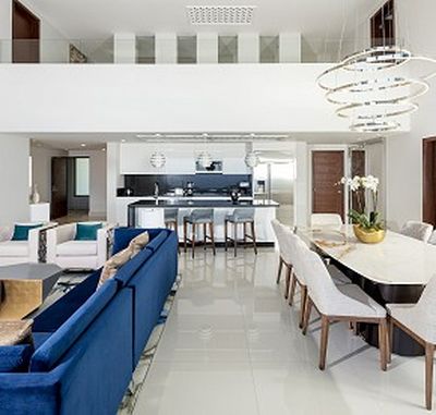 Modern open-plan living area with blue sofa, dining table, and sleek kitchen. White walls, glossy floors, and spiral chandelier add elegance.