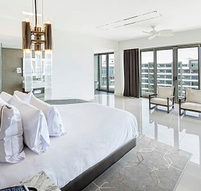 Modern hotel room with a large bed, white linens, a chandelier, two chairs by the window, and a view of a building outside.