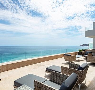 Oceanfront terrace with wicker chairs and glass railing, overlooking a clear blue sea under a partly cloudy sky.