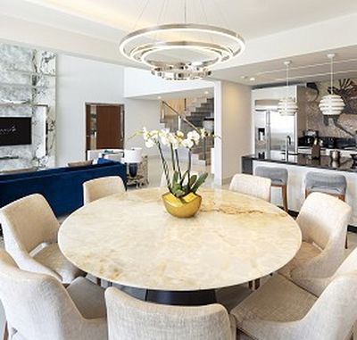 Modern dining area with a round marble table, beige chairs, and an orchid centerpiece. Background features a kitchen and contemporary decor.