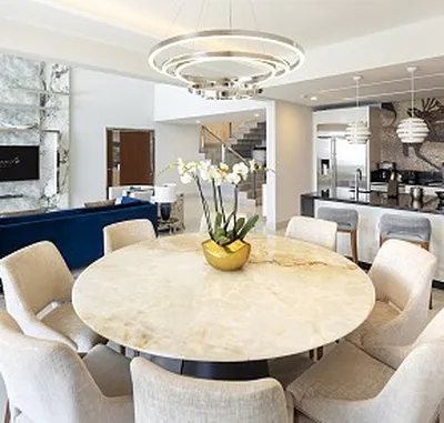 Modern dining area with a round marble table, beige chairs, and an orchid centerpiece. Background features a kitchen and contemporary decor.