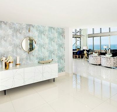Modern living room with white flooring, a patterned wall with a round mirror, sleek furniture, and large windows offering an ocean view.